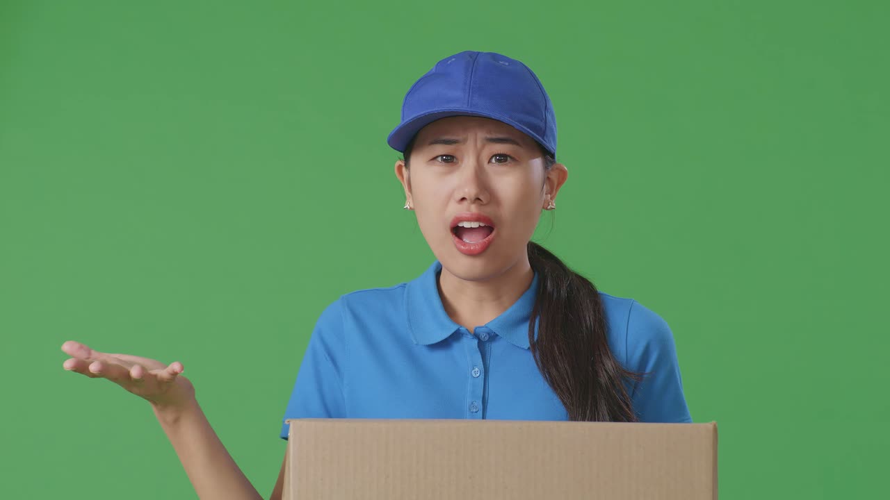 Close Up Of Asian Female Courier In Blue Uniform Showing Puzzled Gesture And Saying What While Delivering A Carton On Green Screen Background In The Studio