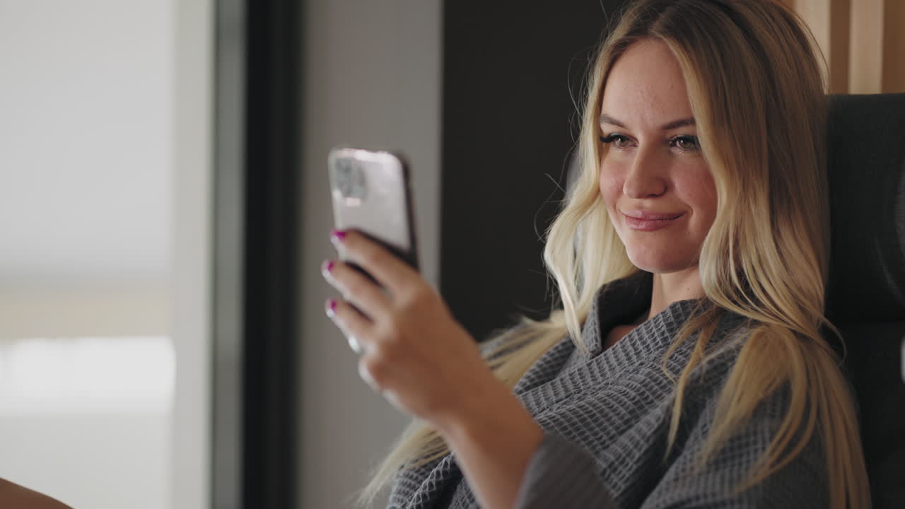 una bonita mujer rubia en bata está usando un teléfono inteligente viendo fotos en las redes sociales descansando en una habitación de hotel o zona de salón