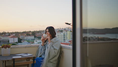 contemplative woman drink coffee on balcony enjoying sunset cityscape alone