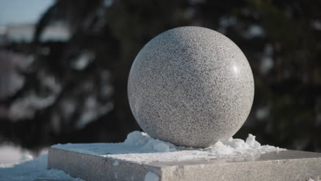 Circular granite sphere sculpture perched on snowy pedestal in urban park under daylight, smooth stone surface reflecting winter light, blurred background of frost coated trees framing seasonal scene