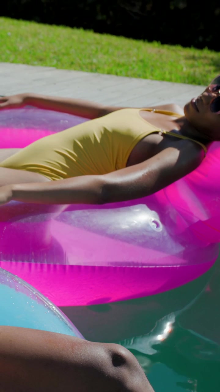 Vertical video: Cam pulling back showing woman in yellow suit on pink float in pool, shades basking