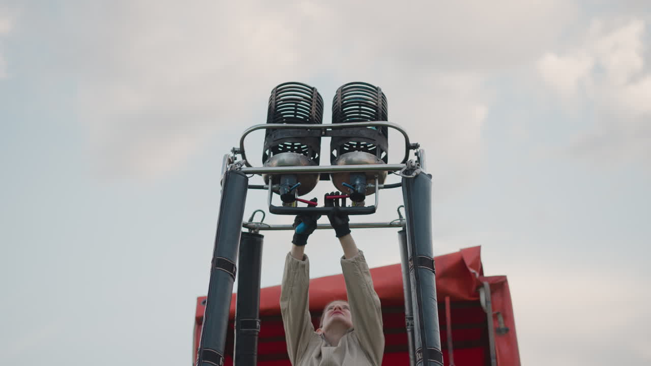 woman extinguishing burner flame on hot balloon frame while securing metal components over grassy field under cloudy sky at sunset with red envelope in background
