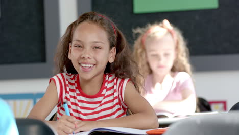 caucasian girl smiles at the camera, biracial girl focuses on her work in a classroom in school