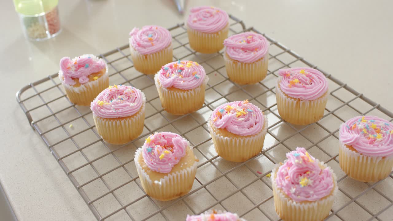 A câmera faz um movimento panorâmico sobre cupcakes em uma prateleira no balcão, mostrando saco de confeitar e granulados para decoração.