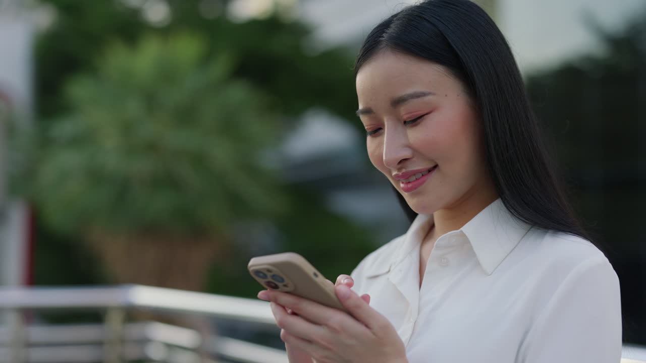 Young Asian woman smiling while using her smartphone outdoors