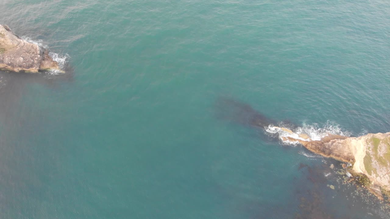 An aerial backward reveal footage of the Lulworth Cove with sandy beach, crystal blue water, fishing boat, kayakistes and crashing waves on the rocky cliff