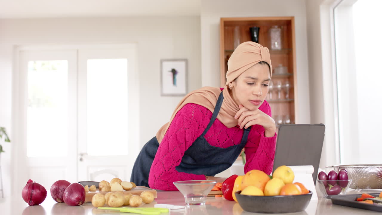 Biracial woman in hijab using tablet preparing food in kitchen at home with copy space, slow motion