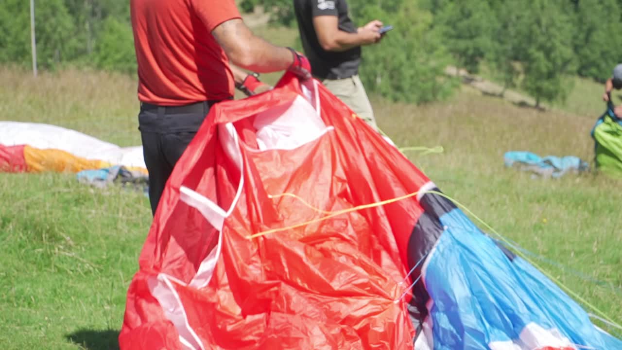 Un parapendista stende con cura la sua vela rossa a terra, preparandosi al decollo, mentre altri fanno lo stesso sullo sfondo.