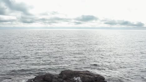 aerial drone view flying over small dark rock island with ice in lake superior revealing waves on water to the horizon on cloudy winter day