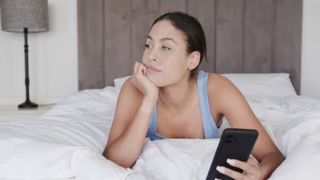 Happy-biracial-woman-lying-on-bed-using-smartphone,-slow-motion