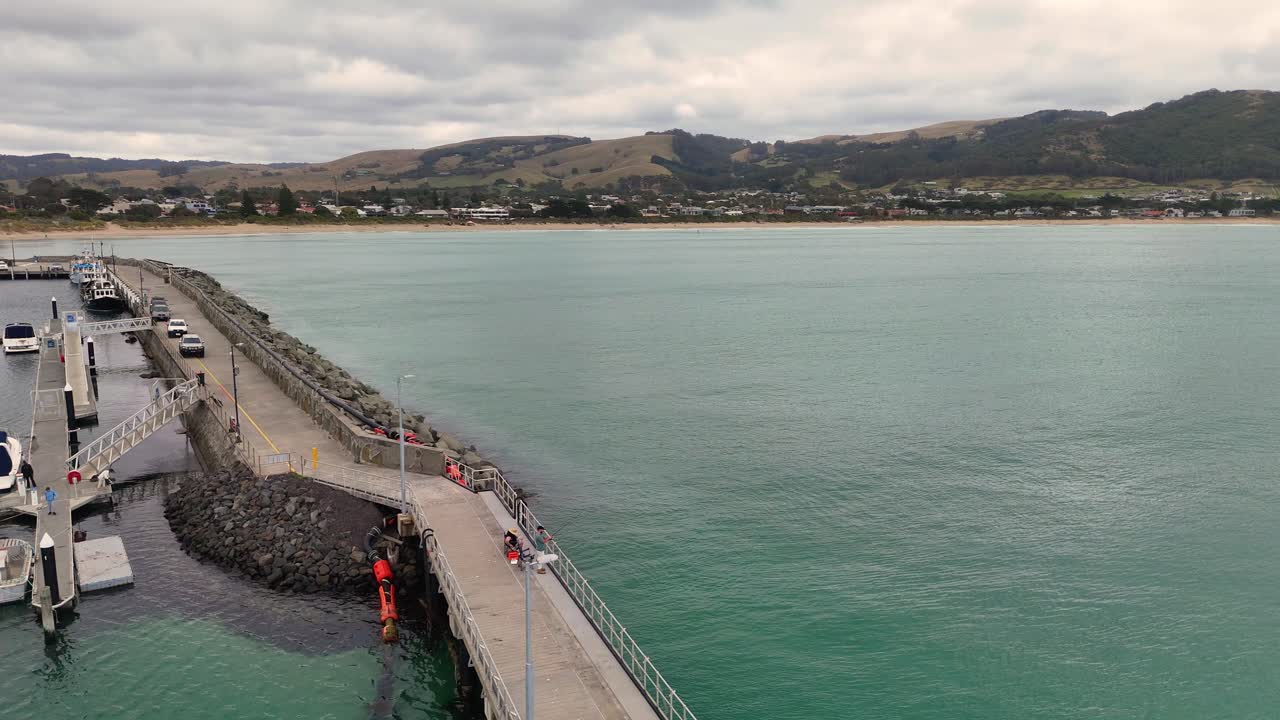 Drone footage captures Apollo Bay pier, boats, and scenic coastline under overcast skies. Calm waters and distant hills create a serene atmosphere