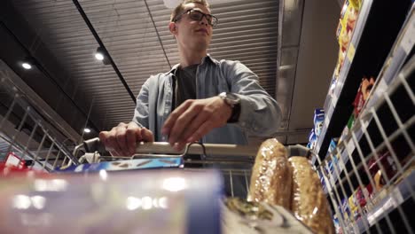 A-cheerful-young-man-in-glasses-putting-goods-in-shopping-trolley.-Passing-through-the-dry-goods-aisle-of-a-big-supermarket.-Low