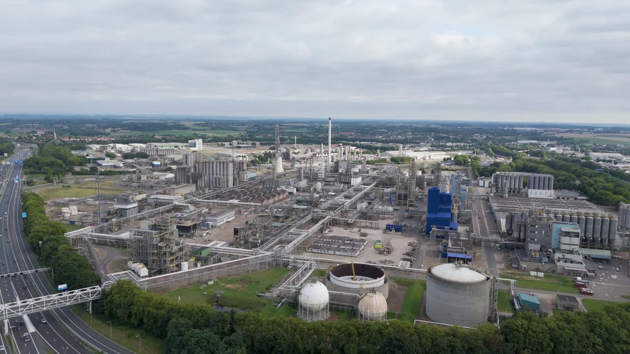 Wide drone footage of a massive industrial refinery with tanks, pipes, and production facilities surrounded by infrastructure