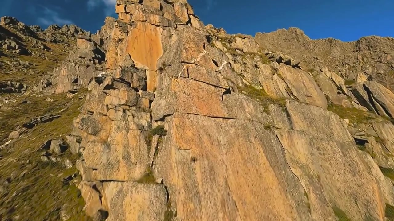 Drone aereo FPV che vola su un crinale montuoso roccioso e ripido con spigoli vivi, durante l'autunno o l'estate. Cielo blu con nuvole bianche sullo sfondo, erba marrone e verde visibile a terra