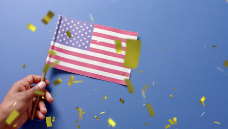 Animation-of-confetti-falling-over-hand-holding-flag-of-united-states-of-america-on-blue-background