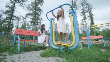 a pregnant mother and her young daughter enjoy playful time together at a playground in the park, surrounded by trees and greenery