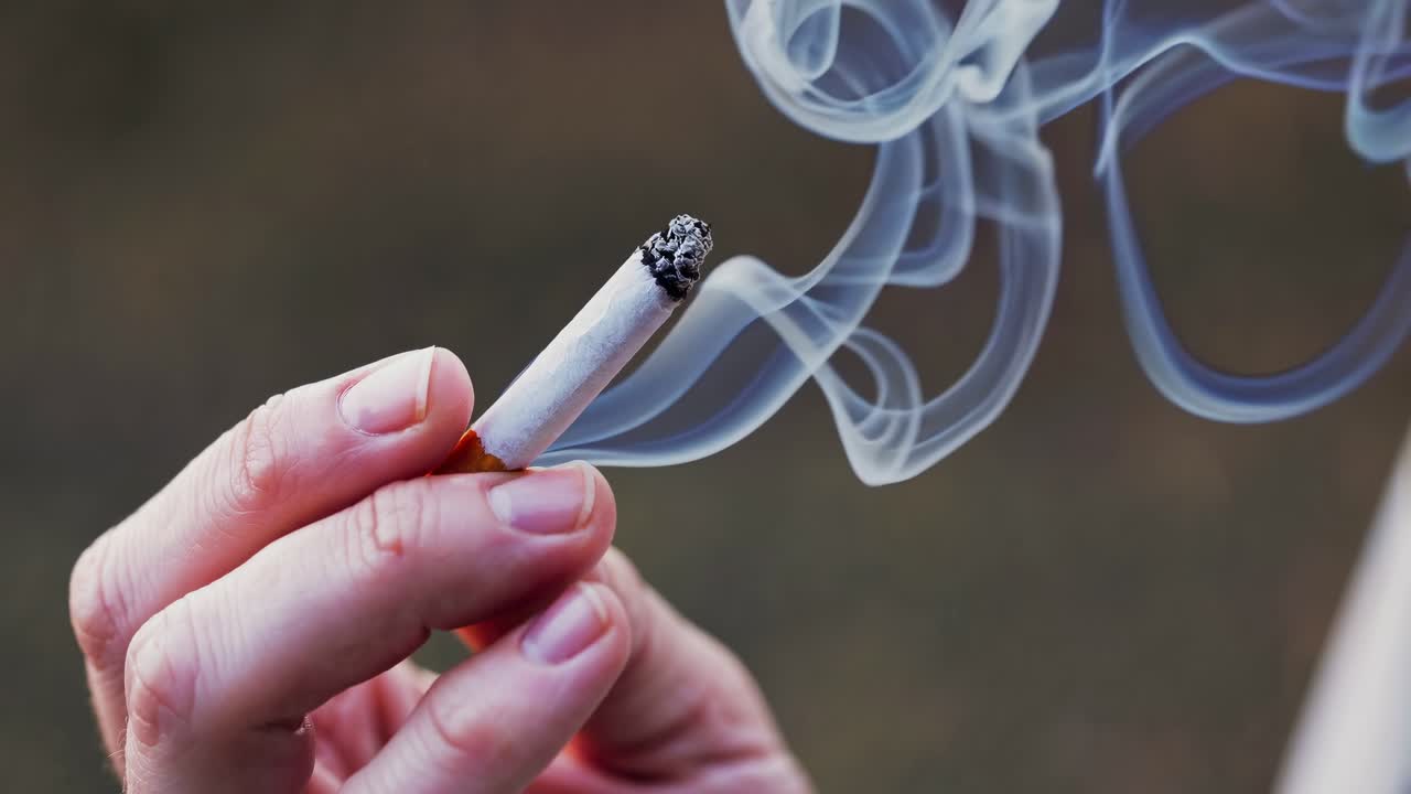Close-up video shot of a hand holding a lit cigarette, with smoke swirling upward