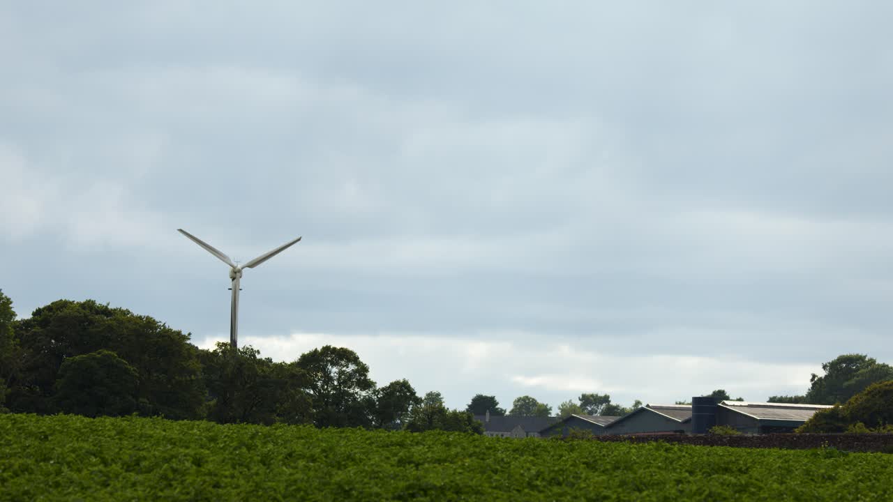 Wind turbine spins steadily above rural landscape under overcast sky, static wide shot, natural lighting