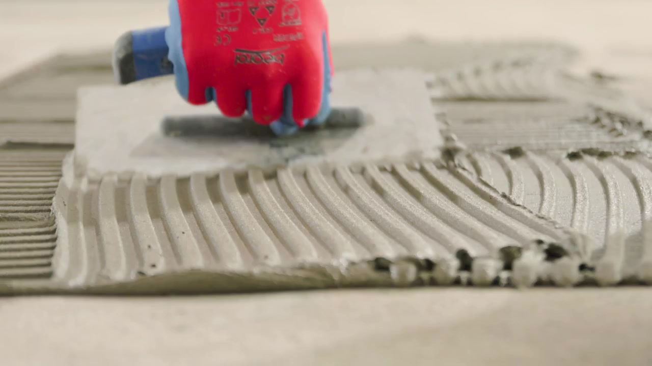 Professional tiler spreading tile adhesive with a notched trowel on the floor of a renovating house.