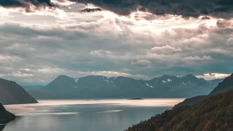 stormy clouds backlit by the setting sun whirl above the calm fjord and mountains in the timelapse video