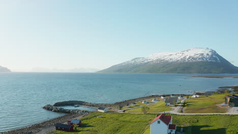 Vista-Aérea-Sobre-Casas-Y-Un-Puerto,-En-Un-Pequeño-Pueblo-En-La-Costa-Del-Mar-De-Barents,-Nevado-Alpino-En-El-Fondo,-Día-Soleado,-En-Troms,-Nordland,-Norte-De-Noruega---Bajo,-Carro,-Tiro-De-Drones