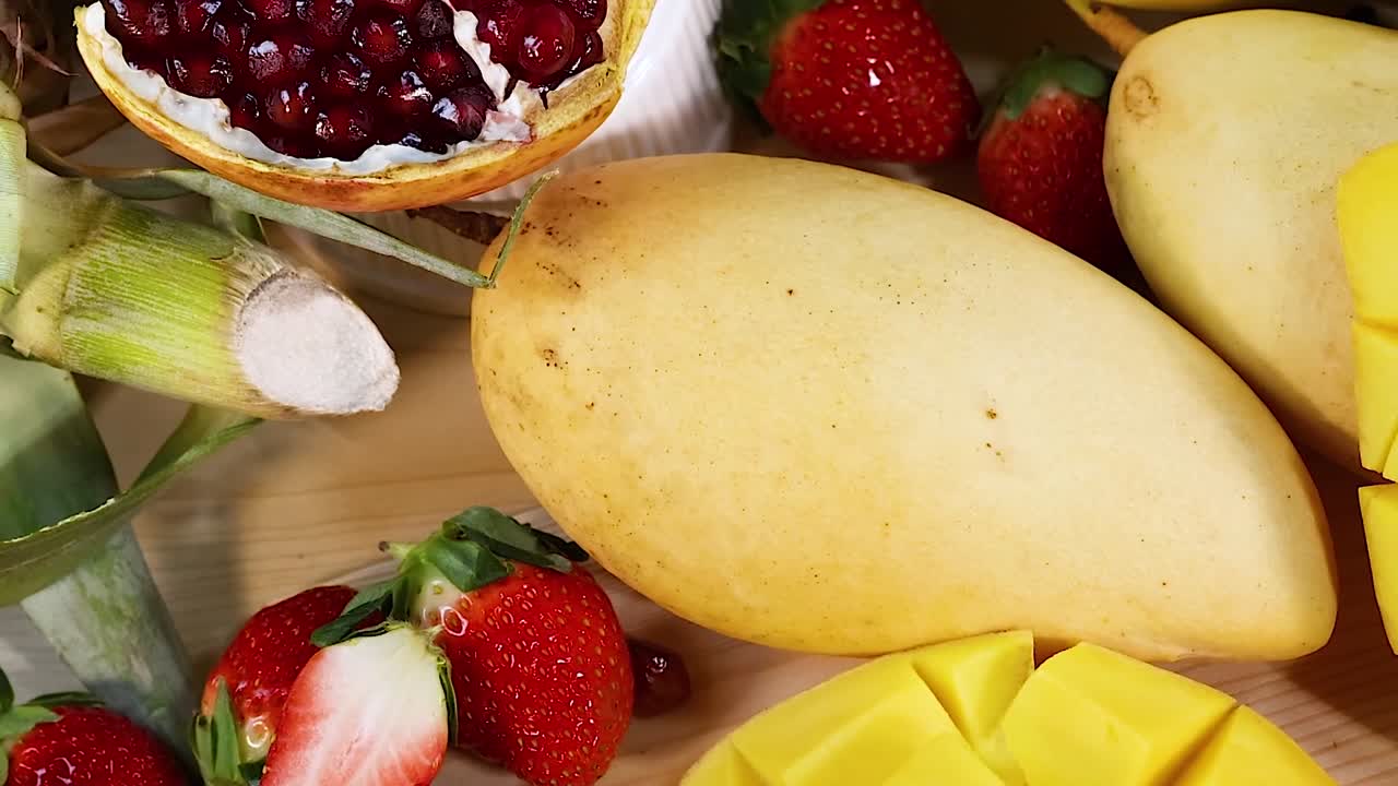 A colorful assortment of sliced mangoes, bananas, kiwis, strawberries, pomegranates, and melons on a wooden surface.