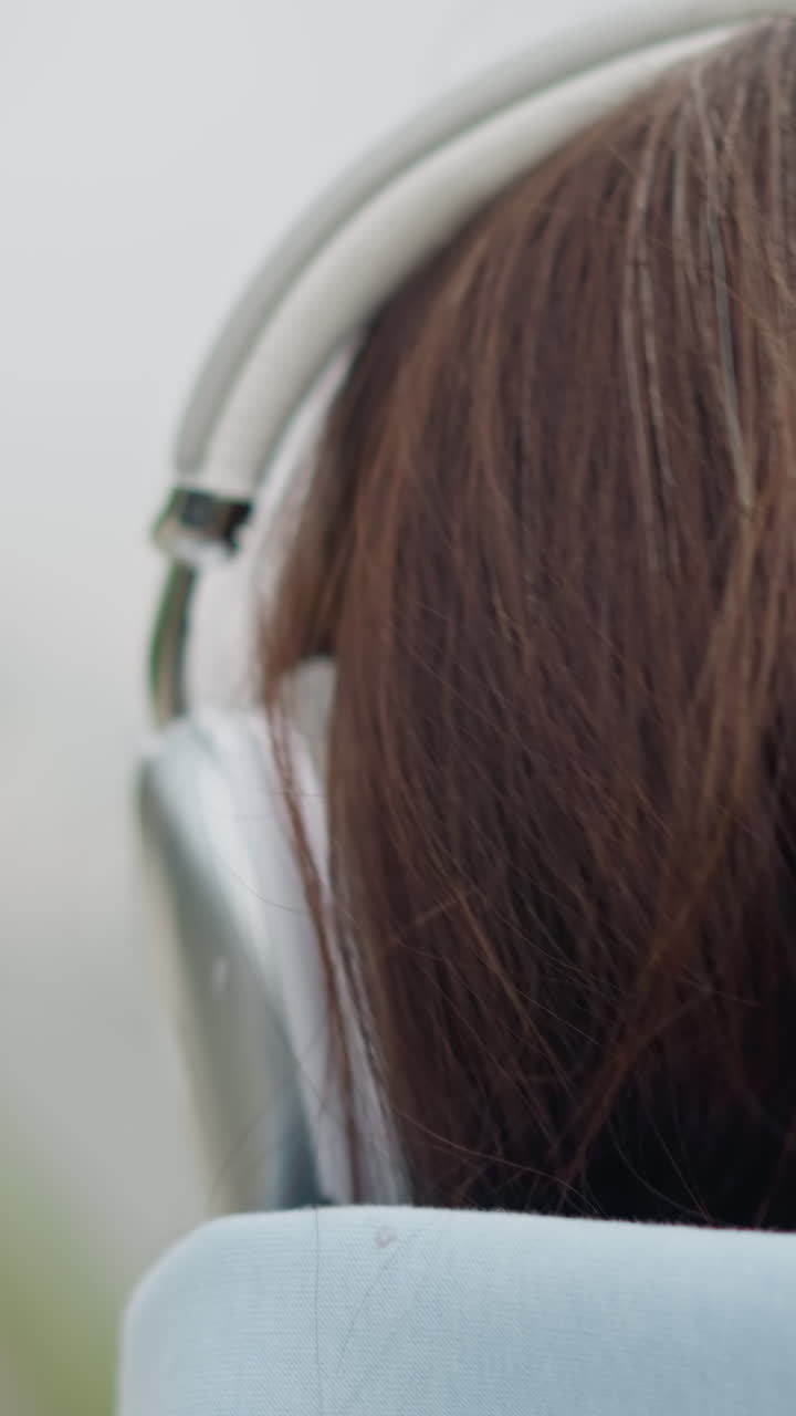 Closeup head view of lady wearing headphones, showcasing her hair and the headset, peaceful outdoor moment, focused expression, enjoying music, urban setting with blurred background