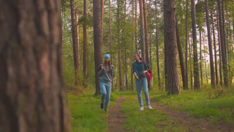 hikers walk along forest trail surrounded by lush greenery, one cousin playfully swings her black bag over her shoulder, while the other, dressed in green shirt, observes with a warm smile