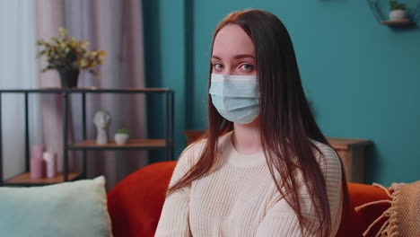 Portrait-of-young-sick-girl-wearing-medical-protection-mask-sitting-in-living-room-looking-at-camera