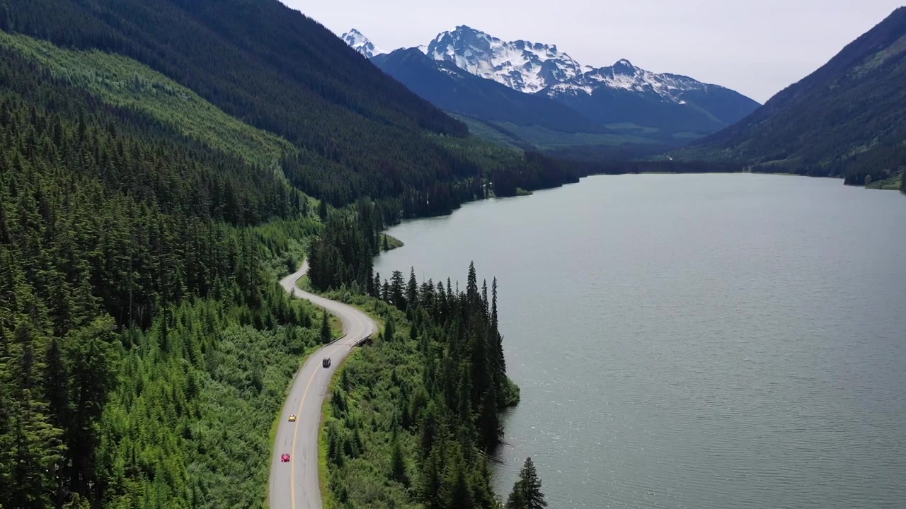 Ripresa aerea con drone dall'alto di un furgone che viaggia su una strada asfaltata attraverso dolci colline e campi verdi. Una scena cinematografica di un viaggio on the road in stile "van life" nell'entroterra della Columbia Britannica sotto un sole splendente nel pomeriggio.