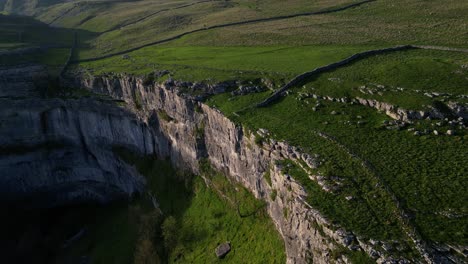 Rising-drone-parallax-around-a-huge-tall-rocky-jagged-cliffside-vertical-drop-at-golden-hour-sunset-surrounded-by-green-hills,-fields-and-countryside-in-yorkshire-england