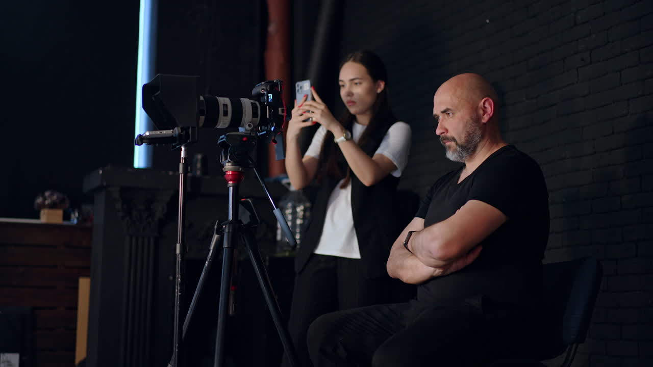 Focused cameraman sits with hands crossed on his chest looking at camera. Brunette girl standing beside takes video on her phone. Photo studio backstage.