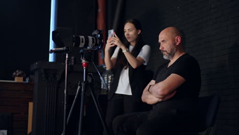 focused cameraman sits with hands crossed on his chest looking at camera. brunette girl standing beside takes video on her phone. photo studio backstage.