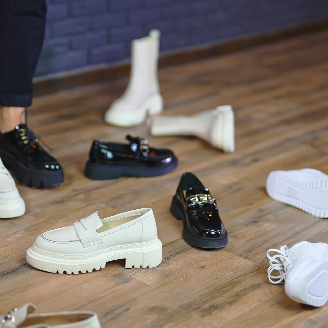 Woman walks in black shoes with tractor soles among other footwear. Black and white footgear scattered on the wooden floor