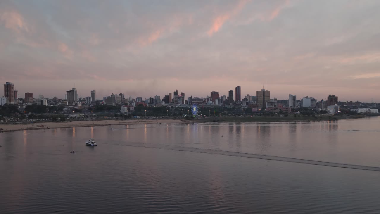 Pan right across the bay of Asuncion during sunset, showcasing waterfront skyline, reflections, and soft pink sky