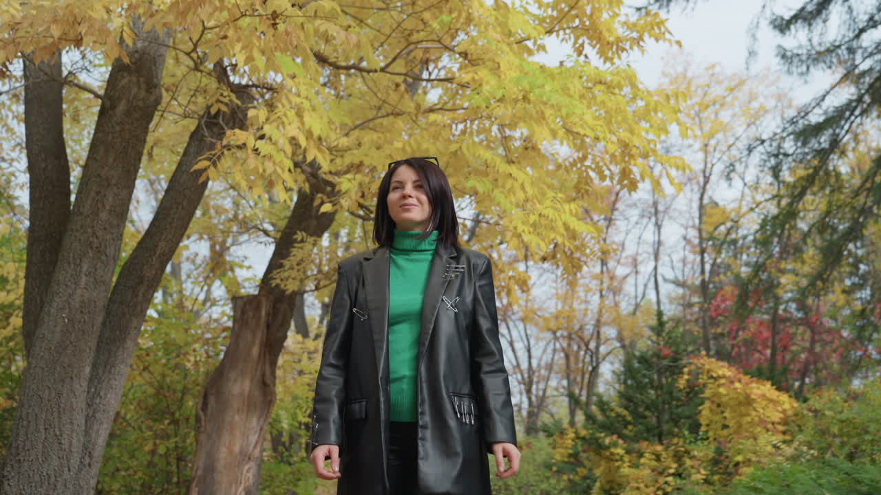 Donna bianca a figura intera con cappotto di pelle in piedi sotto un baldacchino giallo, atteggiamento da rocker sicuro di sé, maglione verde, occhiali da sole pop, stile audace, atmosfera fashion, scatto cinematografico ampio, tranquilla cornice di fogliame autunnale
