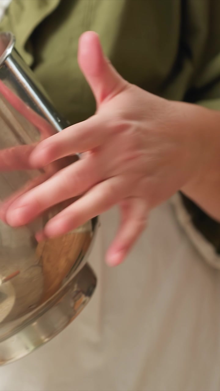 Primo piano di mani che tengono e regolano una ciotola di metallo lucido. La superficie riflettente mostra interazioni tra più coppie. Un indumento verde è parzialmente visibile in alcune inquadrature, suggerendo