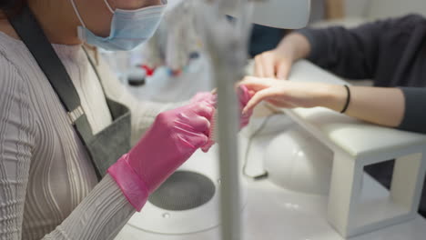 nail technician wearing protective mask and pink gloves carefully grooming customer nails with brush and scissors under bright salon lighting during professional manicure session focused on care