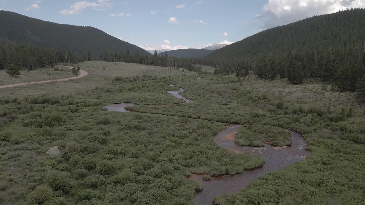 Aerial view of a remote mountain valley with green fields, pine forest and a dirt road. Filmed flying forward in the Rocky Mountains of Colorado near Guanella Pass.