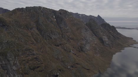 Vista-Aérea-De-Drones-Volando-En-Paralelo-A-Una-Empinada-Pared-De-Montaña-Rocosa-En-El-Nublado-Lofoten,-Noruega