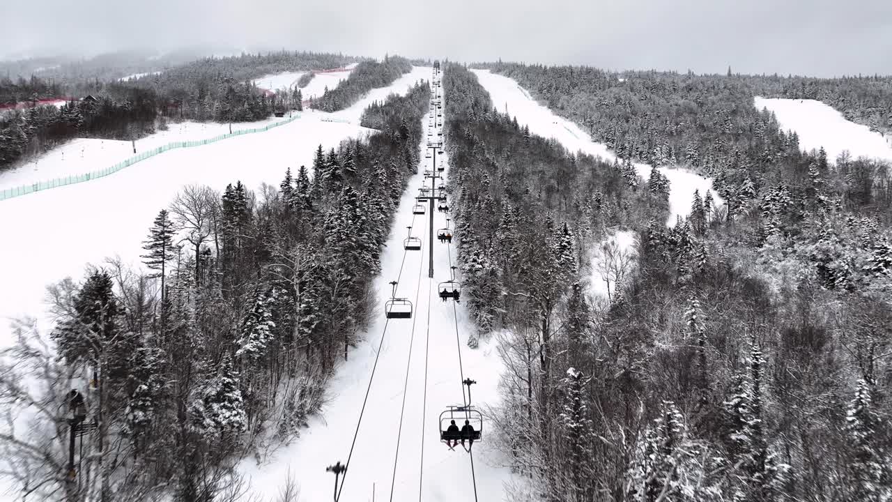 Snowy aerial view of a ski lift ascending a mountain, surrounded by forested slopes covered in fresh powder.
