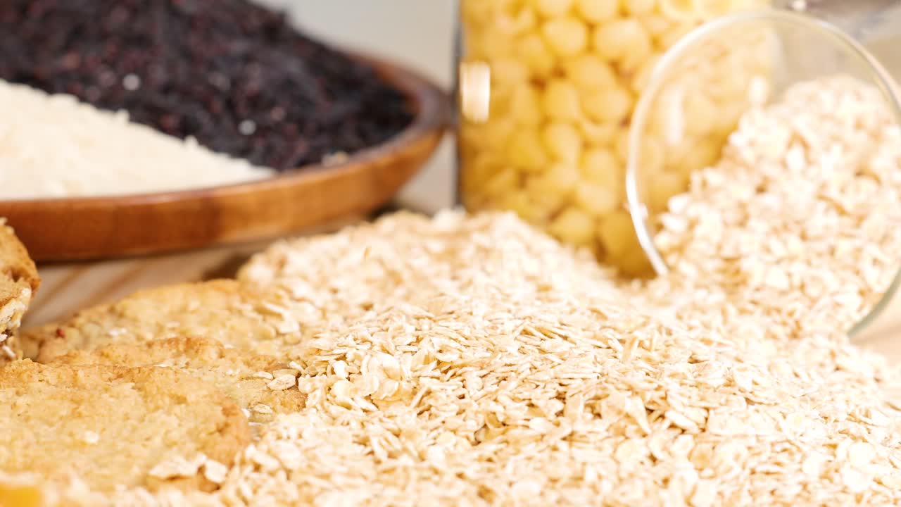 Rolled oats cascade from a container onto a table, surrounded by grains and legumes, in warm lighting