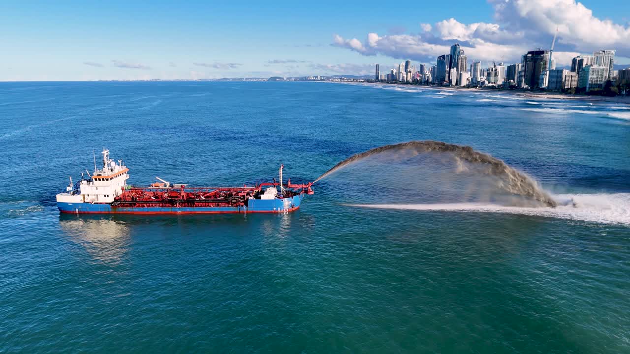A dredging ship pumps sand into the ocean, with a city skyline in the background under clear blue skies