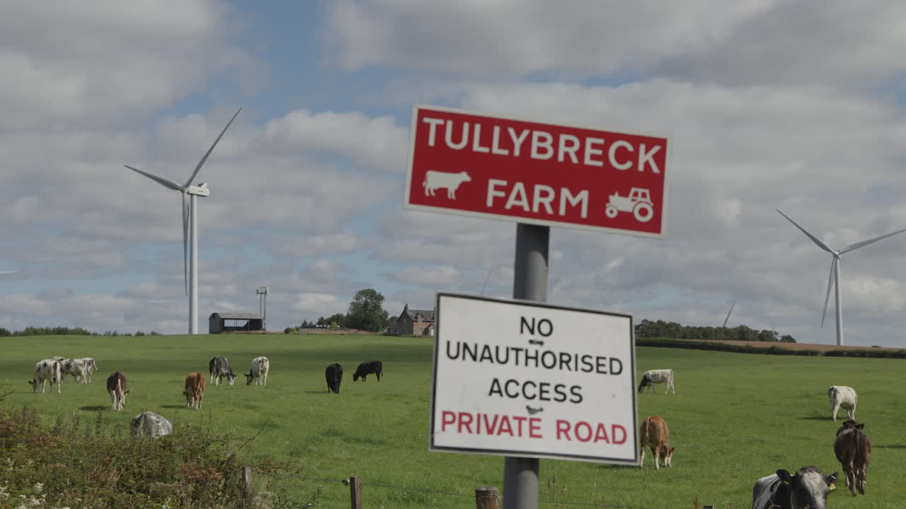 Cows graze in a green field near wind turbines under a partly cloudy sky, with a farm entrance sign and private road warning in the foreground. Static wide shot, natural daylight