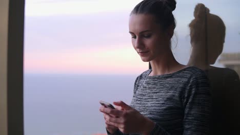 Vista-De-Cerca-De-Una-Hermosa-Joven-Sentada-Junto-A-La-Ventana-Abierta-Con-Una-Sonrisa-Mirando-El-Teléfono-Y-Escribiendo-Un-Mensaje-Durante-El