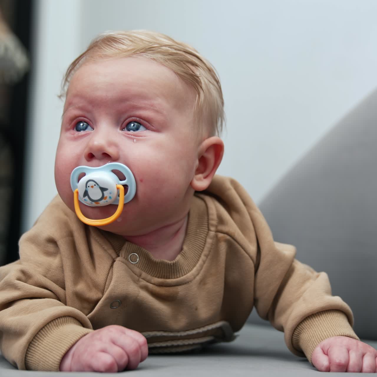 Lovely blond Caucasian baby boy with tears in big blue eyes. Infant child with pacifier lies on belly. Dad's hand wants to get a pacifier but kid turns away. Close up