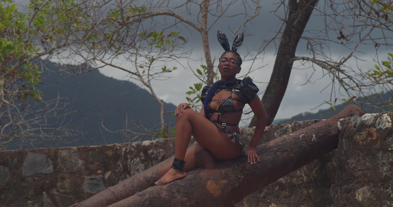 A dark-skinned woman in a luxurious bunny outfit posing atop the weathered stone walls of a Caribbean fort with crashing waves below.