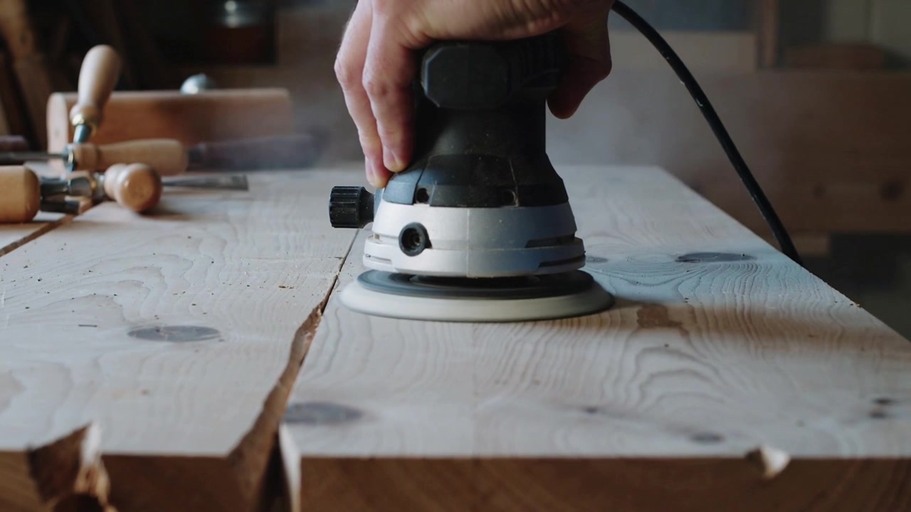 Sanding operator's right hand pressing orbital sander onto seam at workshop bench, smoothing planks