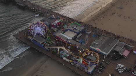 AERIAL:-Circling-Santa-Monica-Pier,-Los-Angeles-from-above-at-beautiful-Sunset-with-Tourists,-Pedestrians-having-fun-at-theme-park-Ferris-Wheel-with-ocean-view-waves-crashing