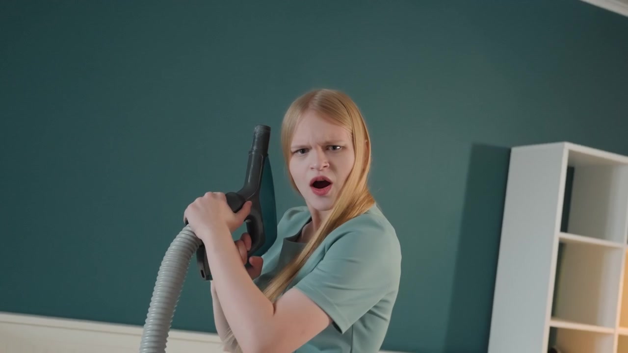 White woman vacuuming house ceiling, determined homeowner pointing vacuum nozzle toward camera, teal wall background, minimal shelf, lowangle framing, focused expressions, cleaning task progression
